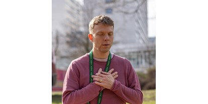 Yogakurs - Yogastil: Yin Yoga - Hamburg-Stadt Farmsen - Ein Mann in meditativer Pose mit geschlossenen Augen und verschränkten Händen über dem Herzen, gekleidet in einen lila Pullover und mit einem grünen Schal um den Hals, der die Ruhe und Zentrierung einer Yin Yoga-Praxis ausstrahlt. Der Hintergrund mit unscharfen Gebäuden suggeriert, dass die Praxis in einer städtischen Umgebung stattfindet. - SOMATiC YiNYOGA