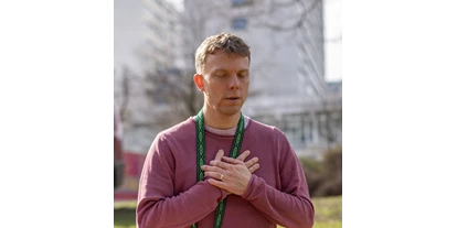 Yoga course - Art der Yogakurse: Geschlossene Kurse (kein späterer Einstieg möglich) - Hamburg-Stadt Grindel - Ein Mann in meditativer Pose mit geschlossenen Augen und verschränkten Händen über dem Herzen, gekleidet in einen lila Pullover und mit einem grünen Schal um den Hals, der die Ruhe und Zentrierung einer Yin Yoga-Praxis ausstrahlt. Der Hintergrund mit unscharfen Gebäuden suggeriert, dass die Praxis in einer städtischen Umgebung stattfindet. - SOMATiC YiNYOGA
